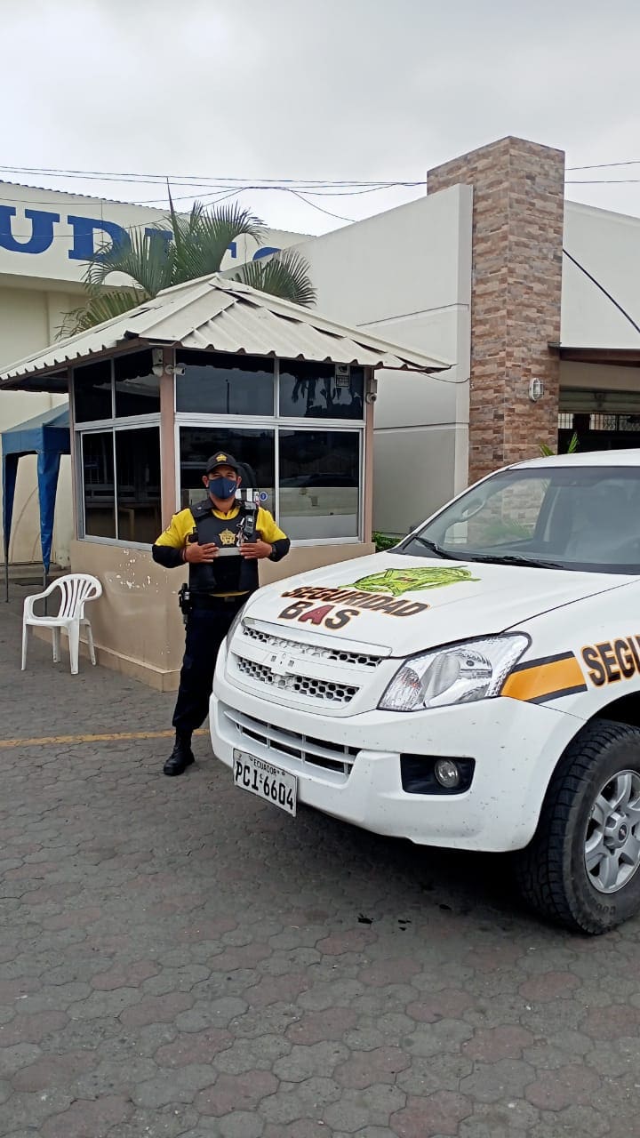 Guardia de Seguridad BAS con chaleco antibalas junto a una camioneta patrullera blanca de la empresa, frente a la garita de control de una planta industrial