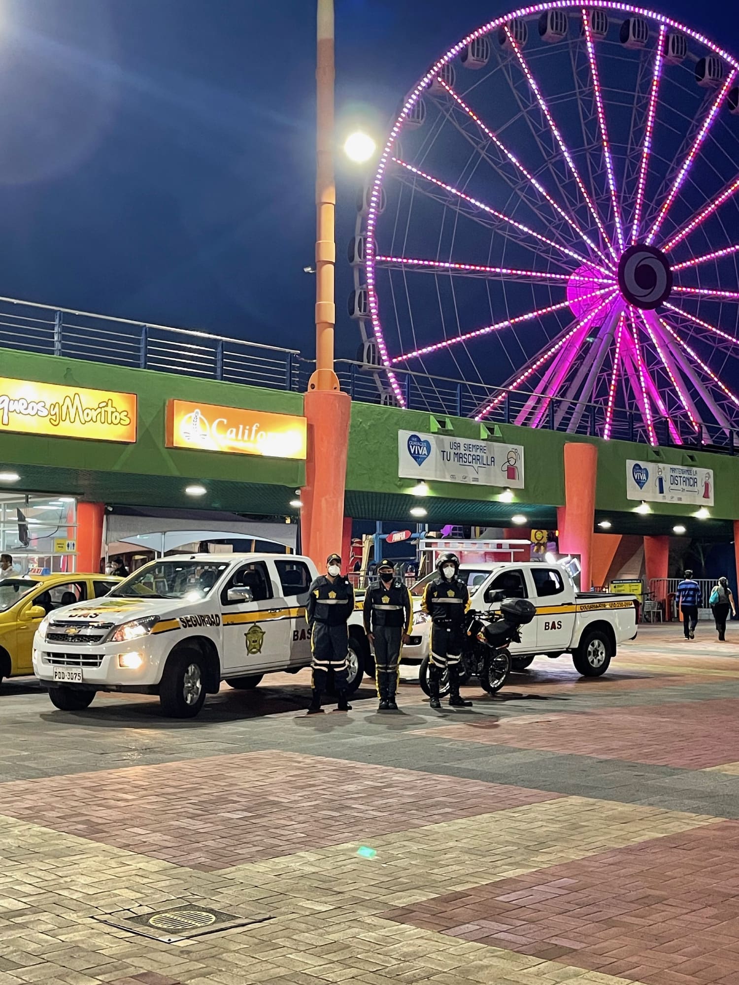 Camionetas de patrullaje y personal motorizado de Seguridad BAS en el acceso de un complejo comercial y de entretenimiento, de noche.