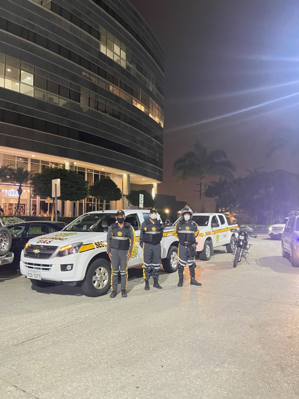 Tres guardias de Seguridad BAS uniformados, de noche, frente a dos camionetas patrulleras blancas de la empresa y una motocicleta, a la entrada de un edificio corporativo iluminado